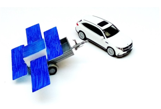 Picture of a cube with solarpanels attached to it. It sits on a trailer, next to the trailer is a car.